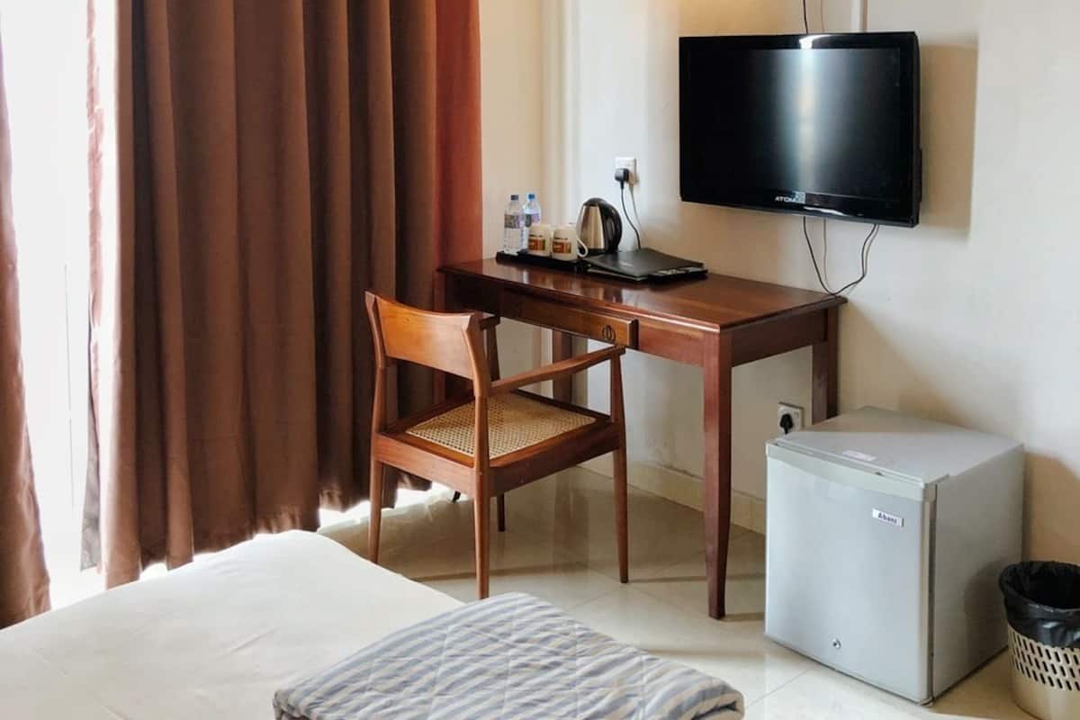 Quarto de hotel com uma cama, uma escrivaninha com cadeira, televisão, mini-frigorífico e ventoinha no teto.