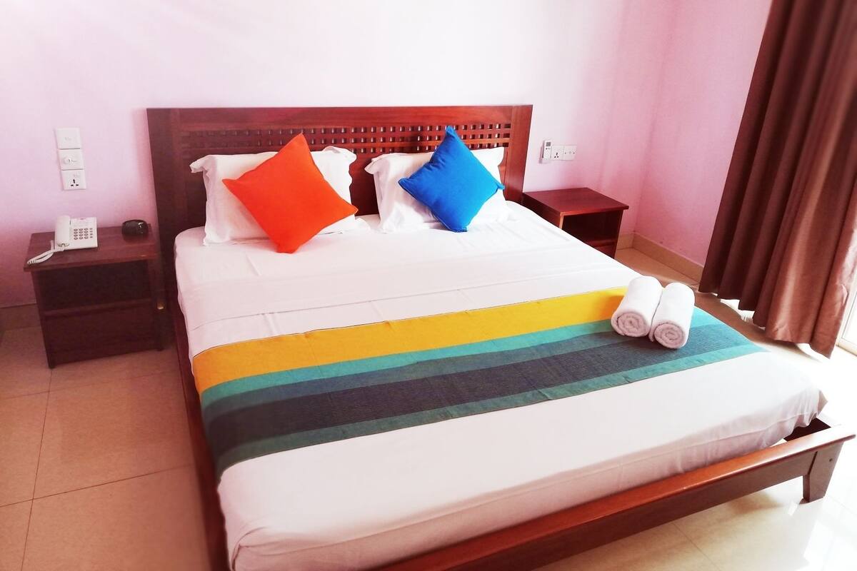 Quarto de hotel com cama de madeira, roupa de cama branca e almofadas coloridas. Há um telefone em uma mesinha de cabeceira e uma toalha enrolada na cama.