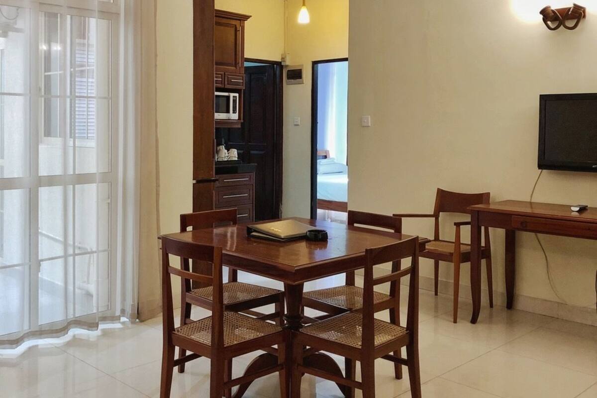 Uma sala de jantar com mesa e cadeiras de madeira, um ventilador de teto e uma cozinha ao fundo.