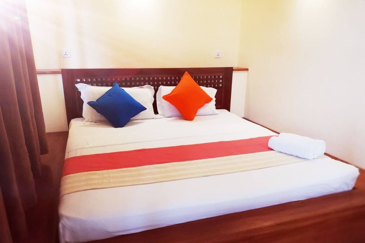 Uma cama bem arrumada com lençóis brancos, um cobertor listrado em vermelho e bege, e dois travesseiros (um azul, um laranja).