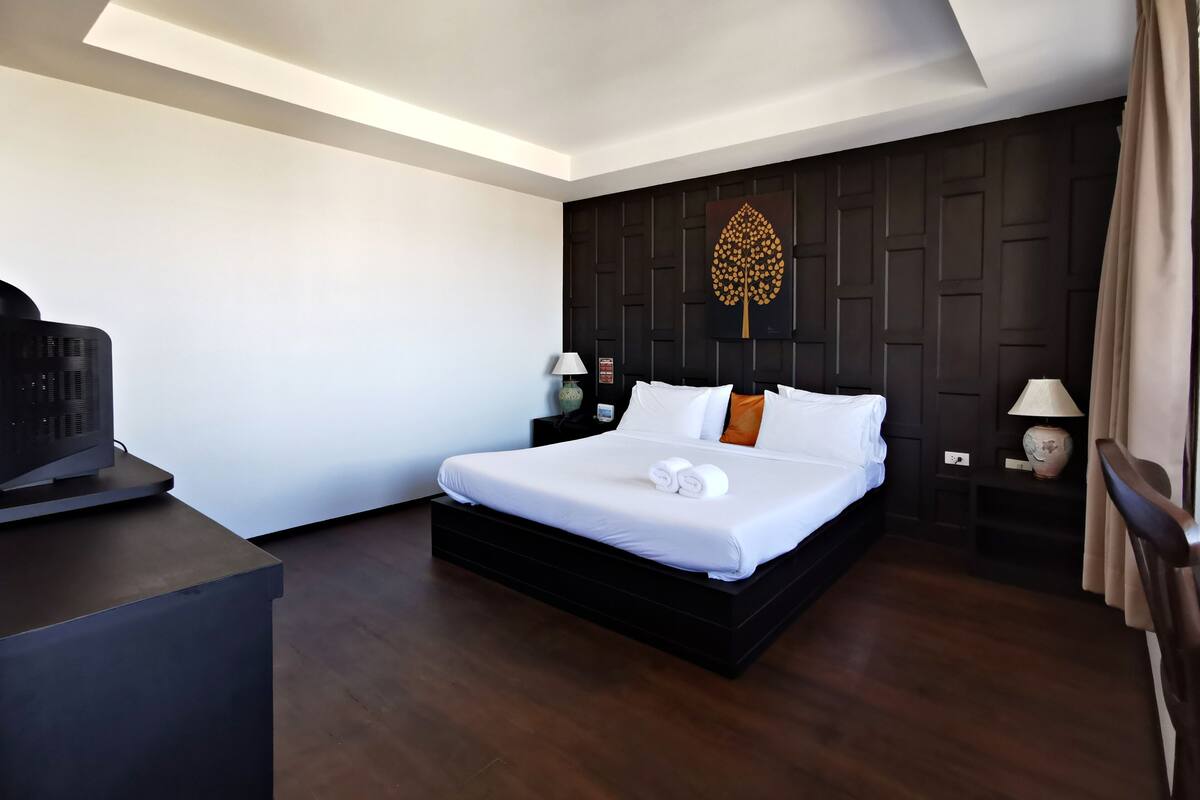 A modern hotel room with a large bed, dark wood flooring, a flat-screen TV, two bedside tables with lamps, and a decorative wall panel.