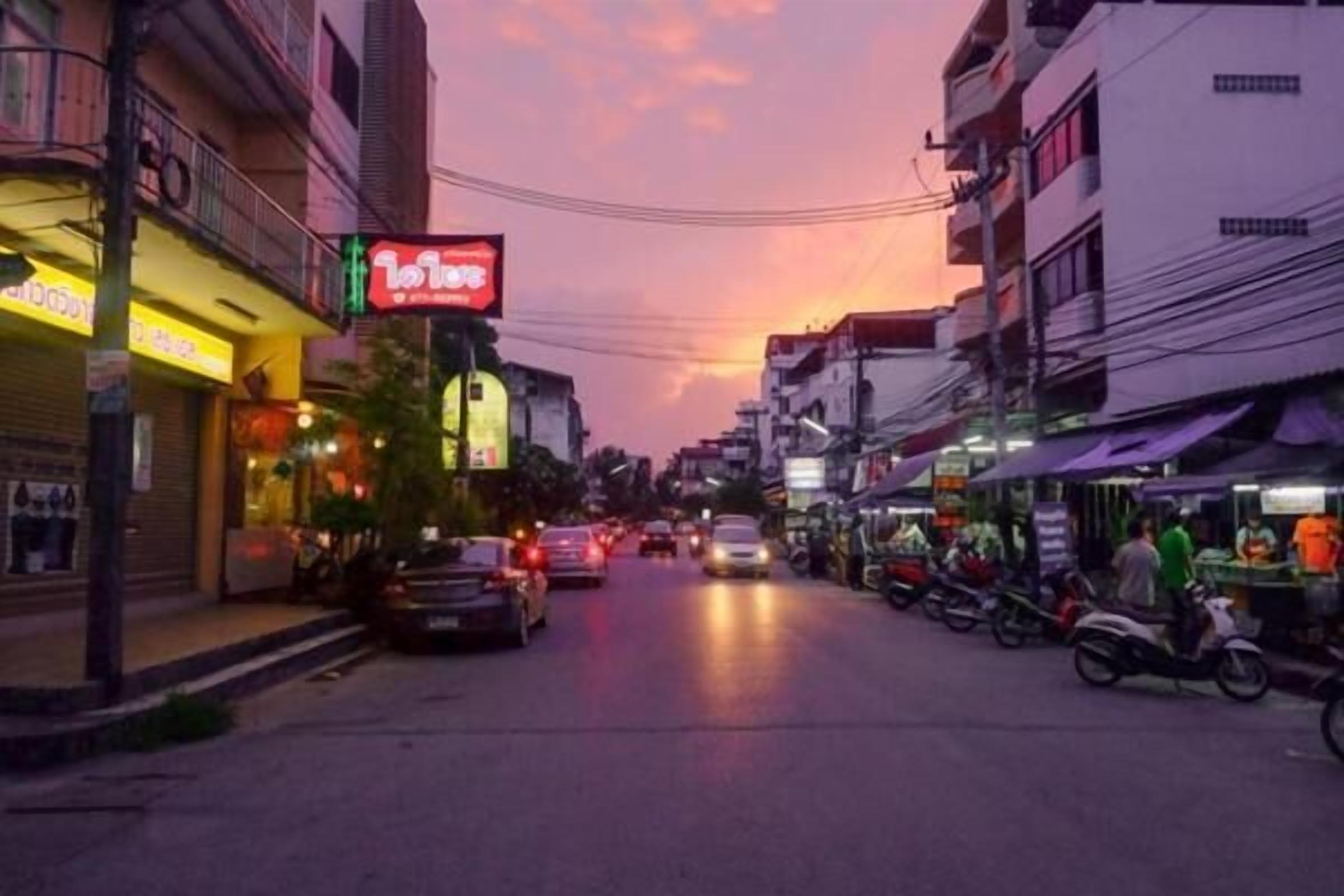 A street scene at dusk with buildings, shops, and parked vehicles.