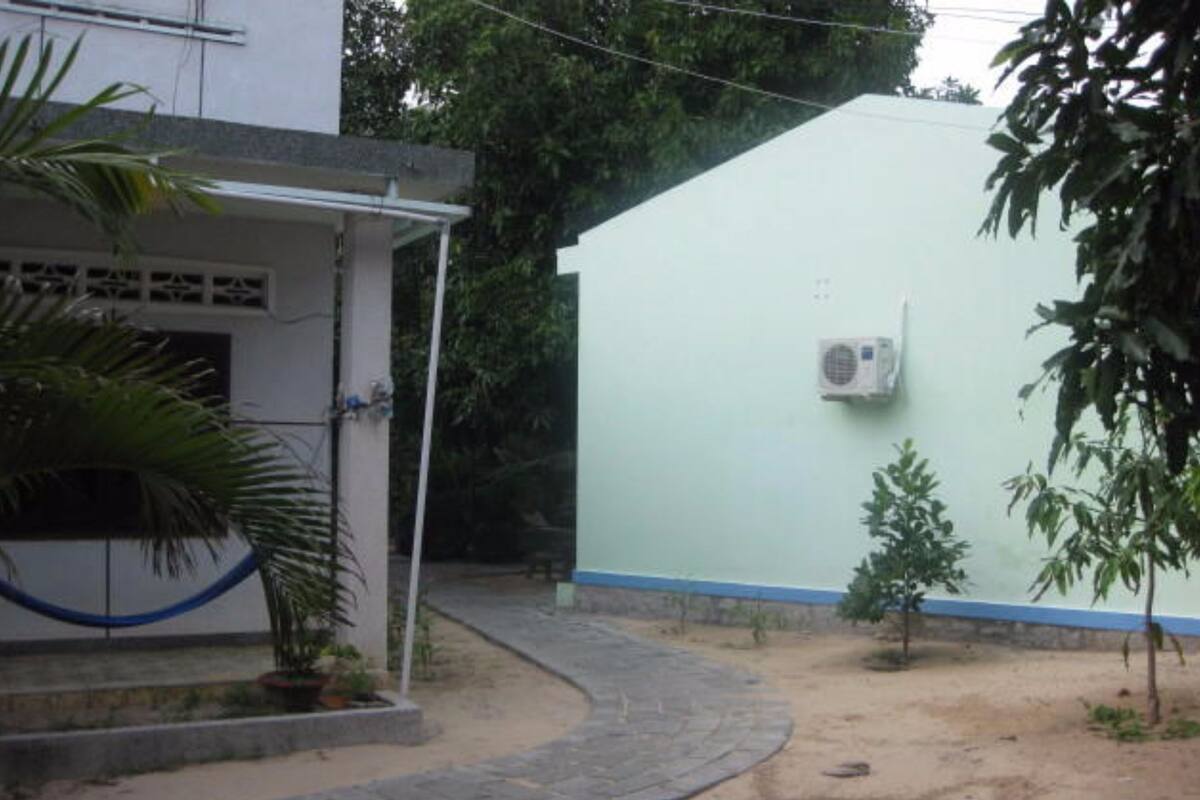Un patio empedrado con un sendero curvo que conduce a un edificio de fachada blanca y detalles azules. Se observa una unidad de aire acondicionado pequeña montada en la pared.