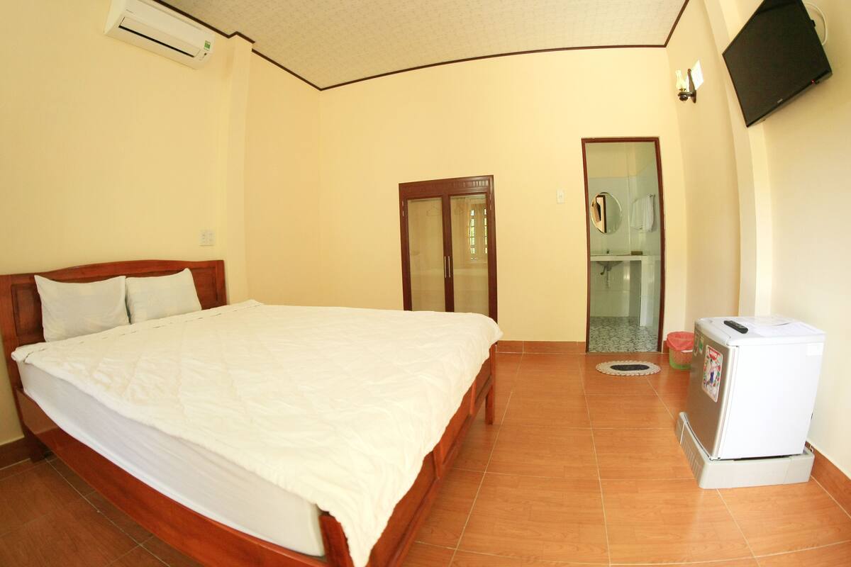 Habitación de hotel con una cama de madera, un refrigerador pequeño y un baño visible a través de una puerta de cristal.