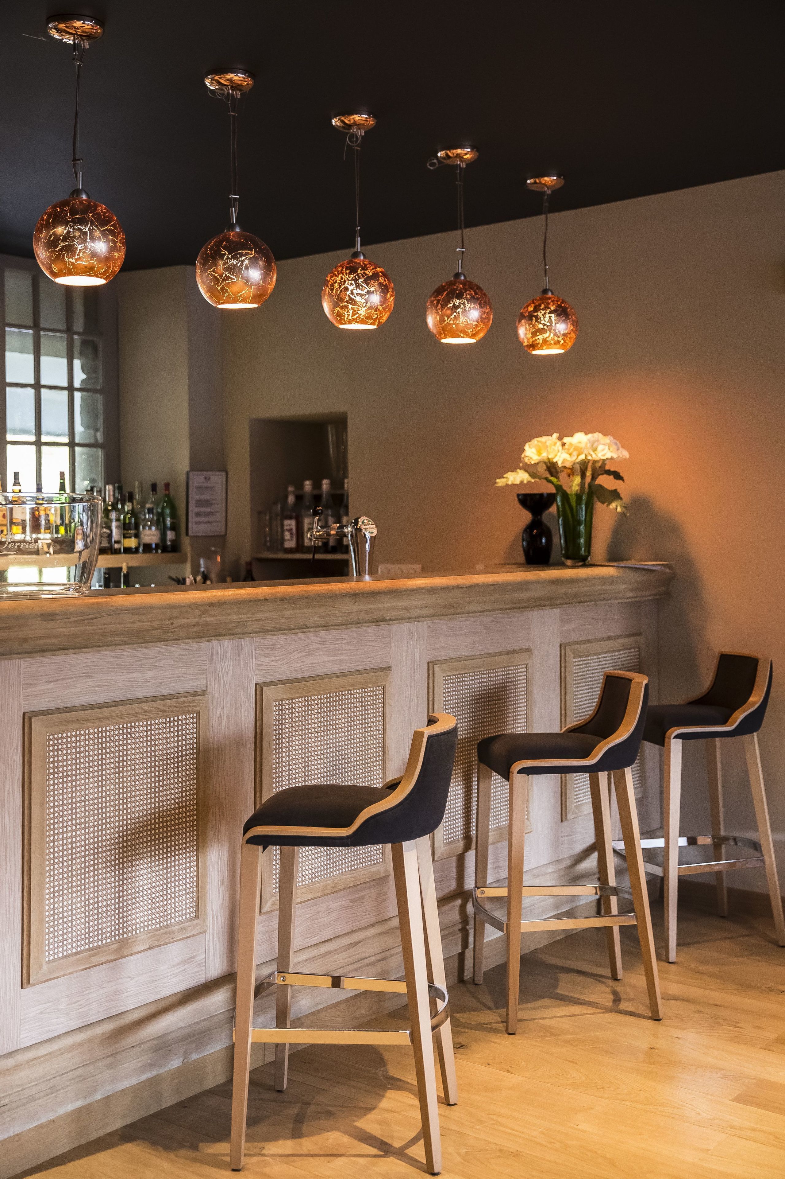 Una zona de bar con taburetes de madera, lámparas colgantes esféricas y una barra con un jarrón de flores.
