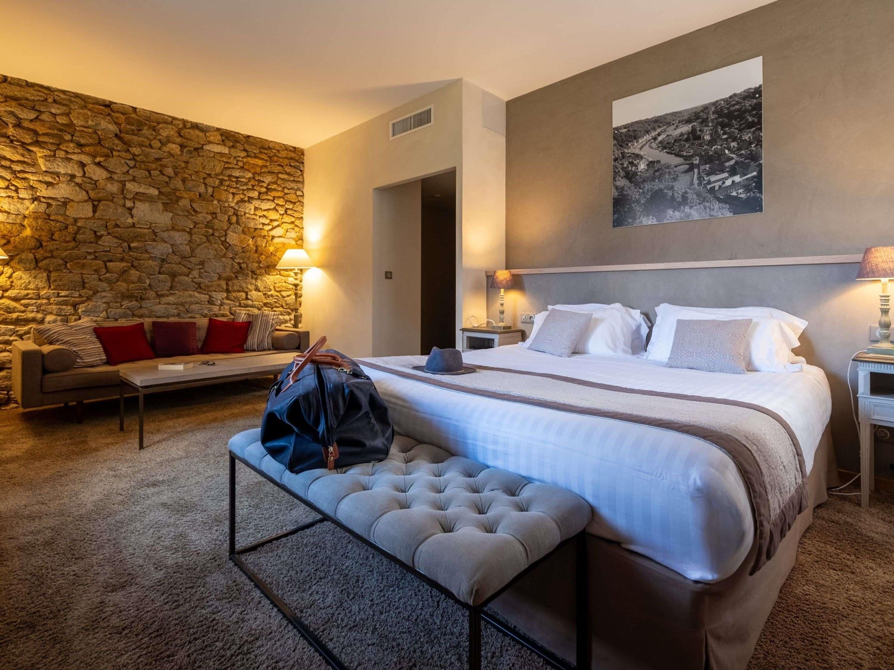 Habitació d'hotel amb paret de pedra, un llit amb roba de llit blanca, una panxa i una tauleta de nit amb una làmpada.