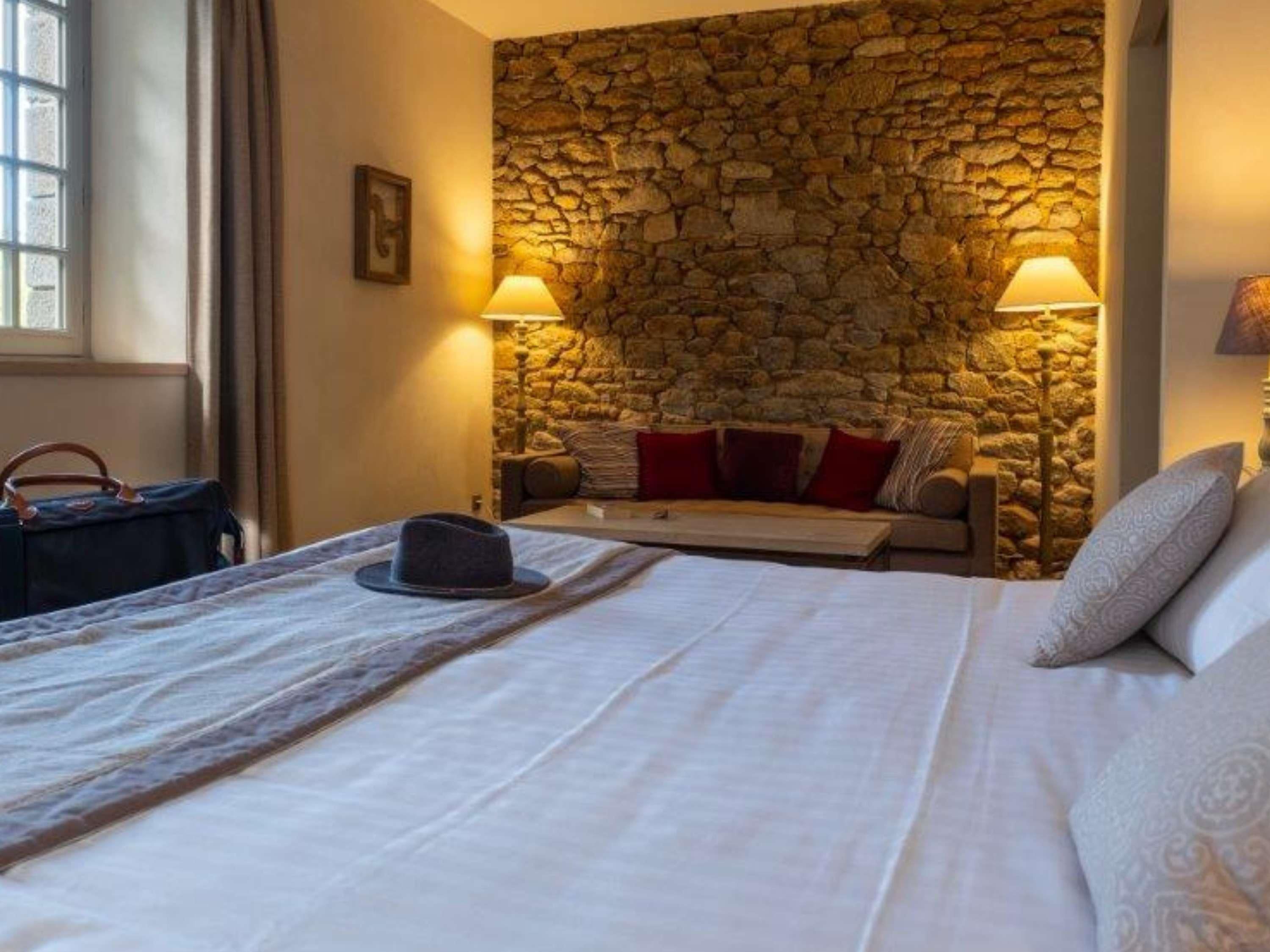 A hotel room with a stone wall, a bed with white linens, a sofa with red cushions, and two lamps.