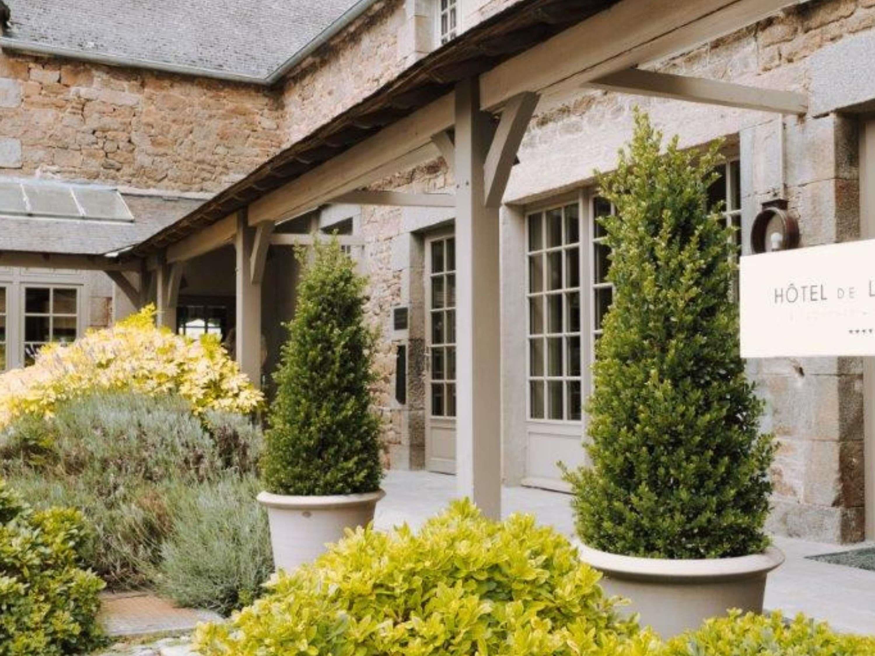 A stone building with a sign reading 'Hôtel de L' indicating a hotel. There are potted plants and a covered walkway.