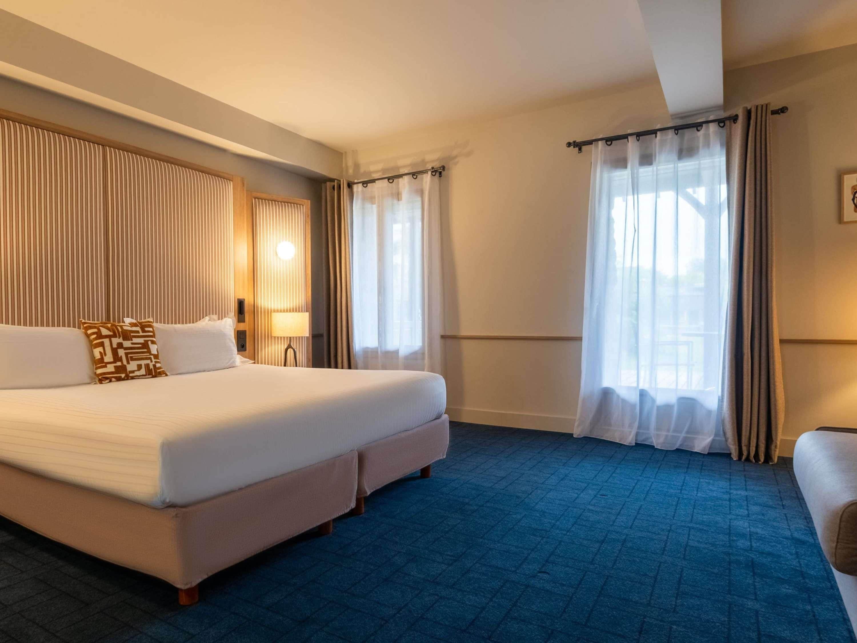Une chambre d’hôtel avec un grand lit, une table de chevet, une lampe et une fenêtre avec des rideaux.