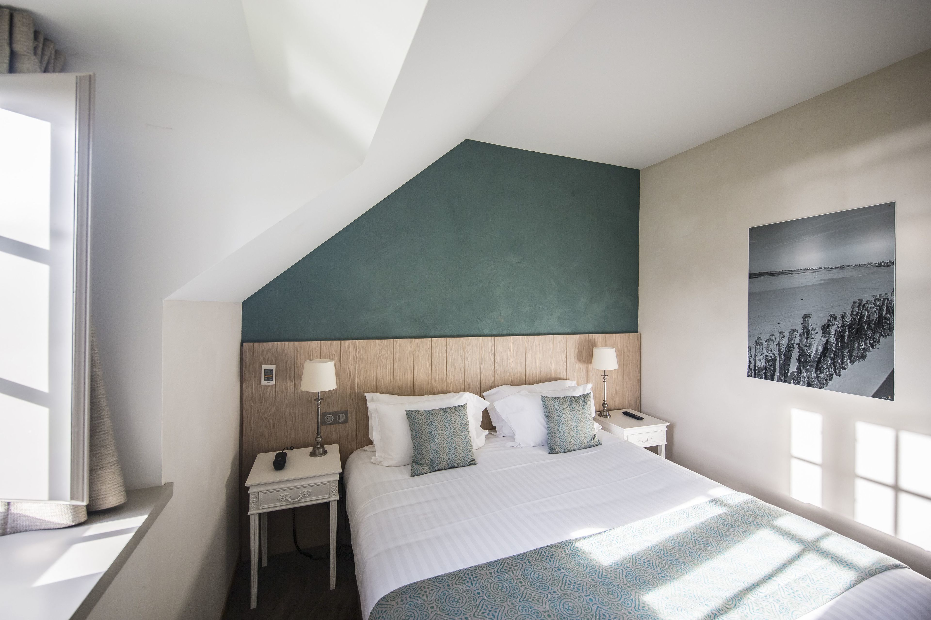 Une chambre d’hôtel moderne avec un plafond en pente, un grand lit avec une literie à motifs, des lampes de chevet, une table de chevet et un tableau accroché au mur.