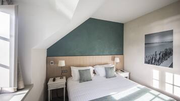 Une chambre d’hôtel moderne avec un plafond en pente, un grand lit avec une literie à motifs, des lampes de chevet, une table de chevet et un tableau accroché au mur.