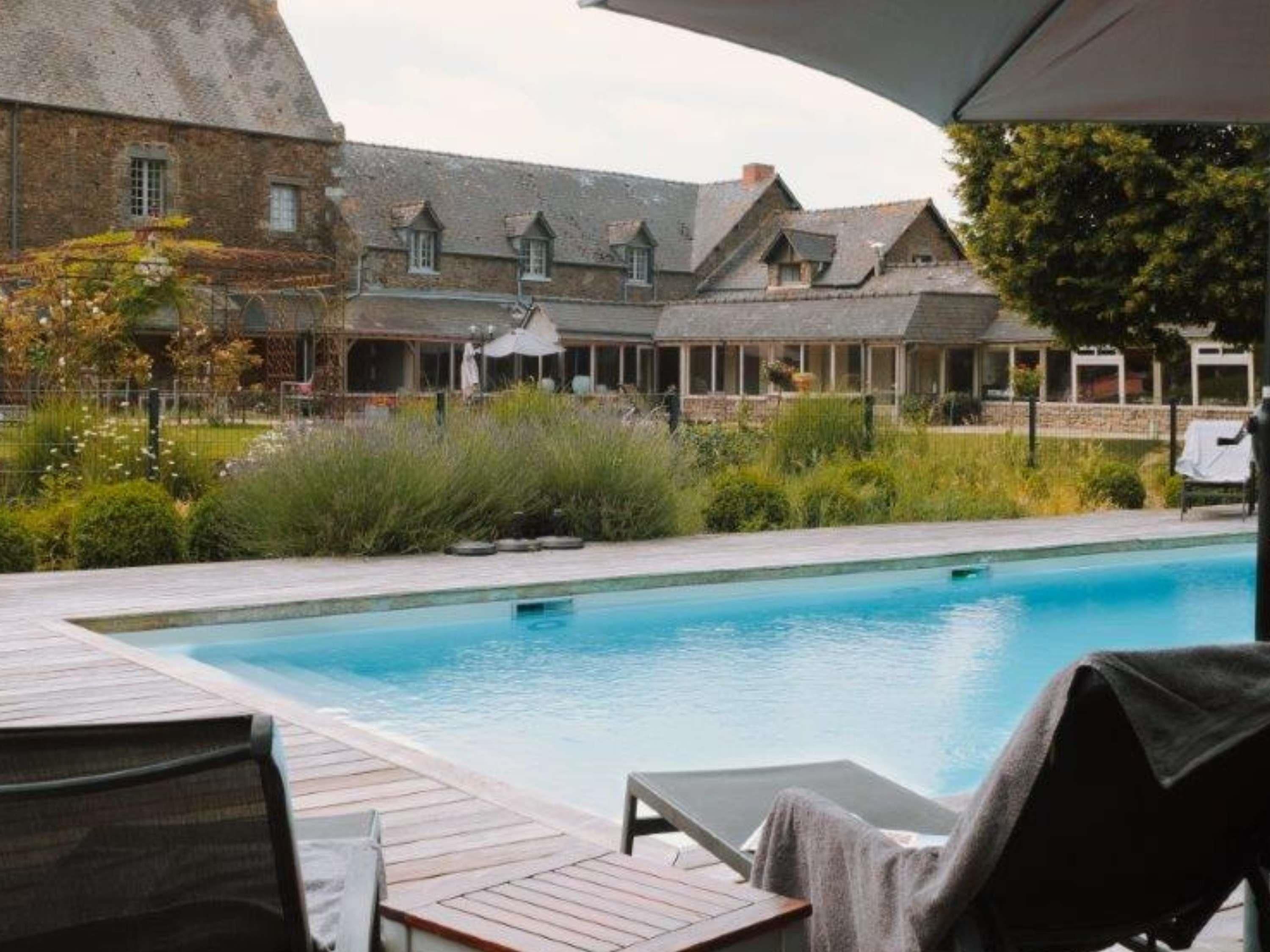 Une piscine dotée de chaises longues et d’un espace abrité, entourée d’un jardin et d’un bâtiment avec de nombreuses fenêtres.