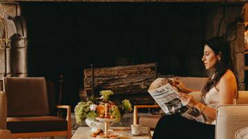 A woman reading a newspaper in a cozy room with a fireplace, a wooden bench, and a table with a floral centerpiece and pastries.