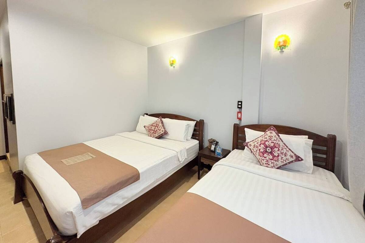 Ein Hotelzimmer mit zwei Einzelbetten, beide mit hölzernen Kopfteilen, weißer Bettwäsche und gemusterten Kissen. Es gibt Nachttische mit Lampen und einen kleinen Beistelltisch.