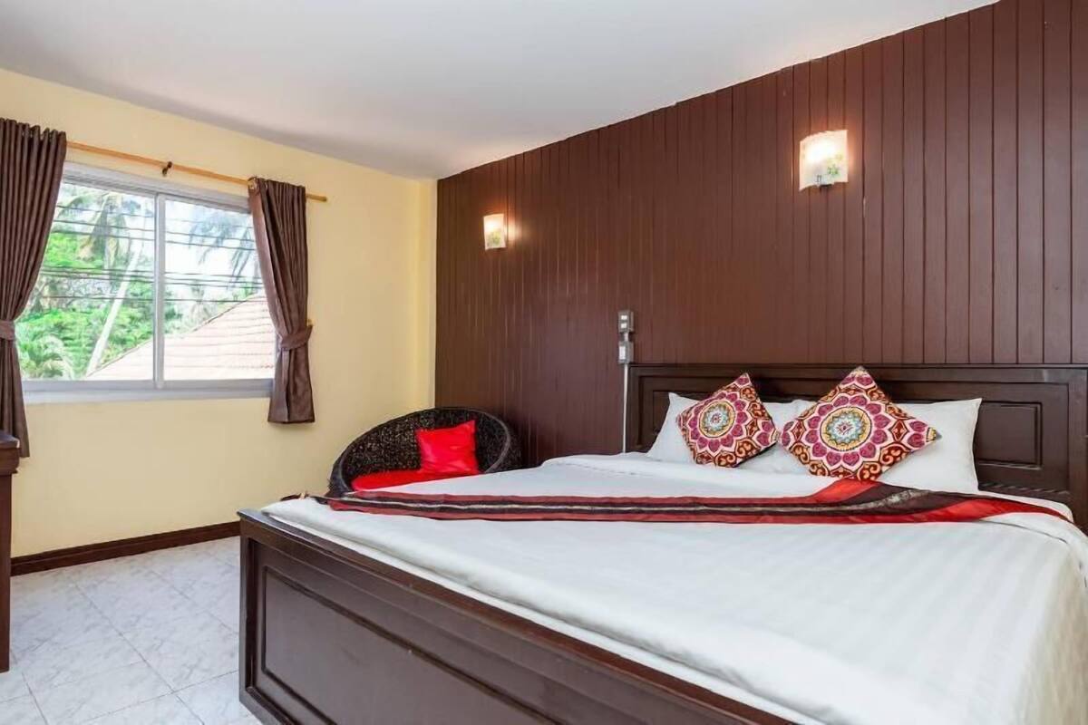 Ein Schlafzimmer mit Bett, Vorhängen, Nachttisch, Wandlampe und einem Fenster mit Blick ins Grüne.