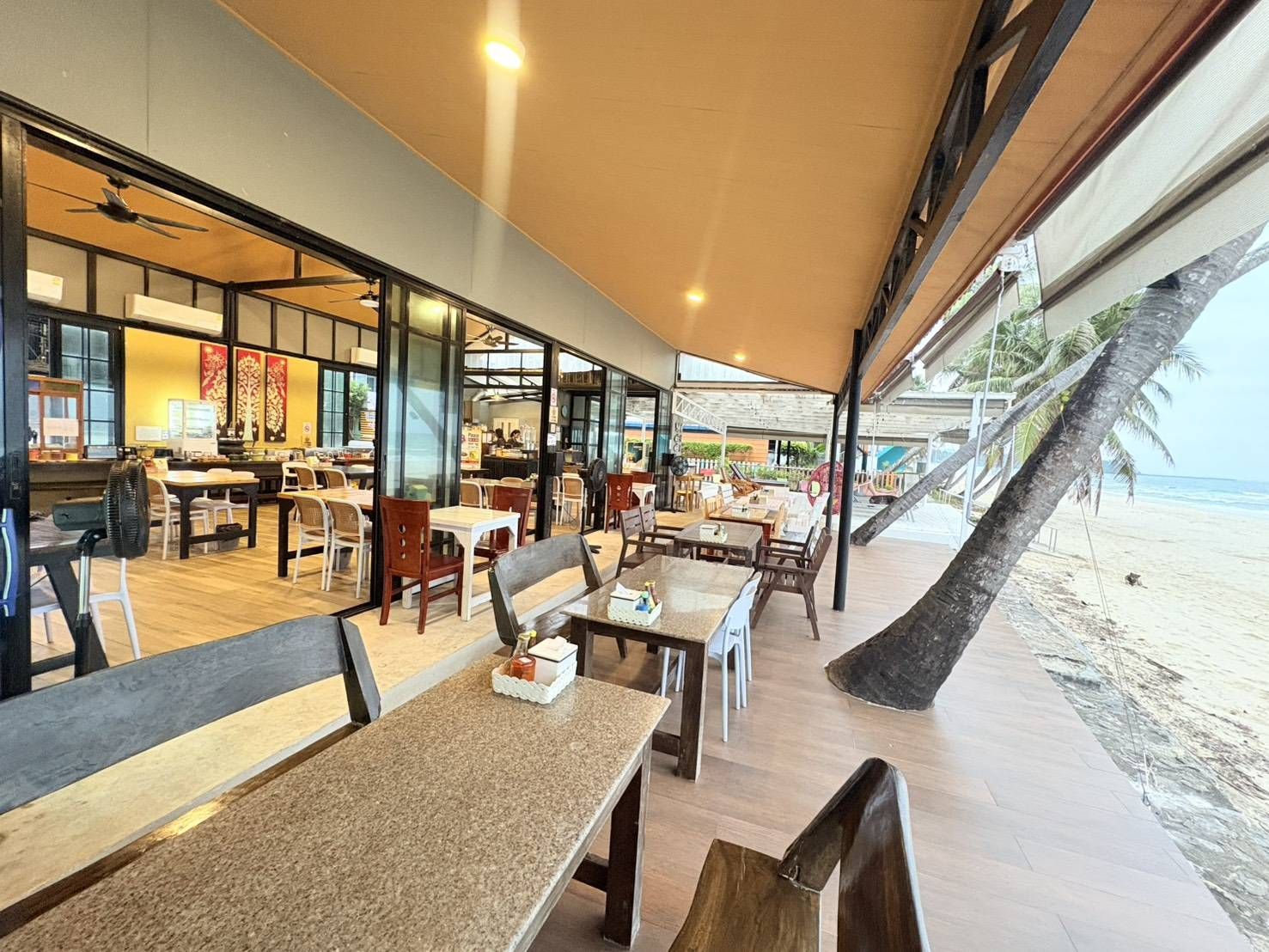An indoor dining area with tables and chairs, a view of the beach, and a palm tree.