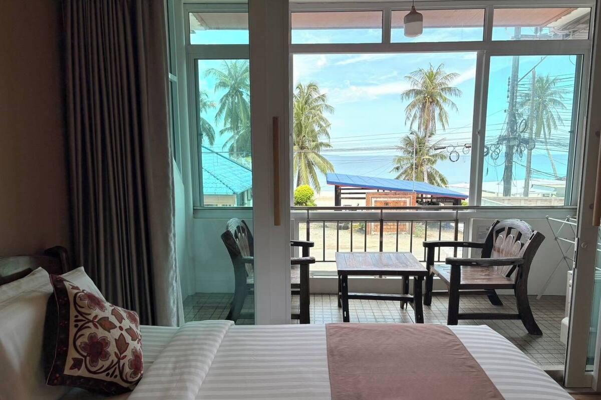 Ein Hotelzimmer mit einem großen Bett, einem Balkon mit Blick auf Palmen und einen Strand, sowie einem kleinen Tisch und Stuhl.