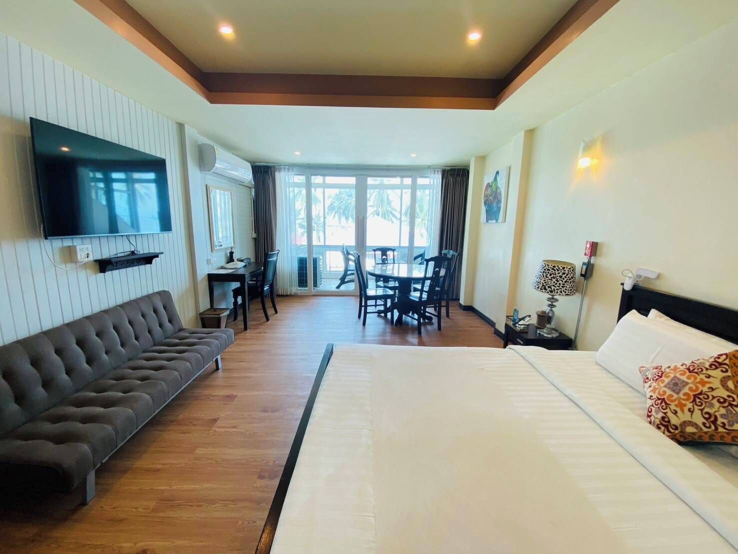 Habitación de hotel con una cama grande, un sofá, un televisor montado en la pared, zona de comedor con mesa y sillas, y balcón con vistas.