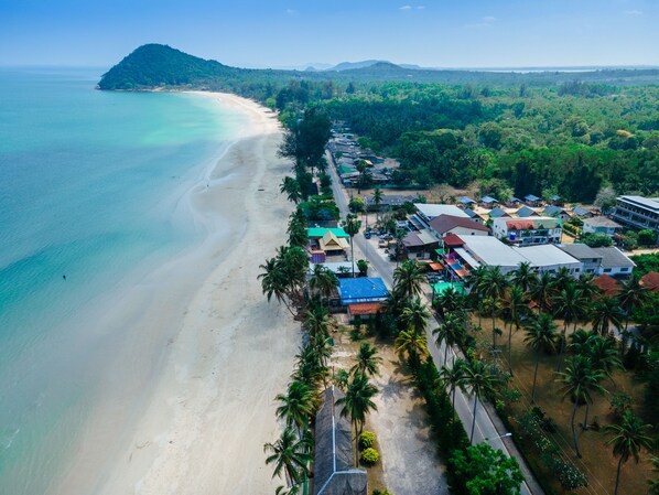 An aerial view of a coastal area with a beach, a small hill, and a cluster of buildings with a road leading to them.