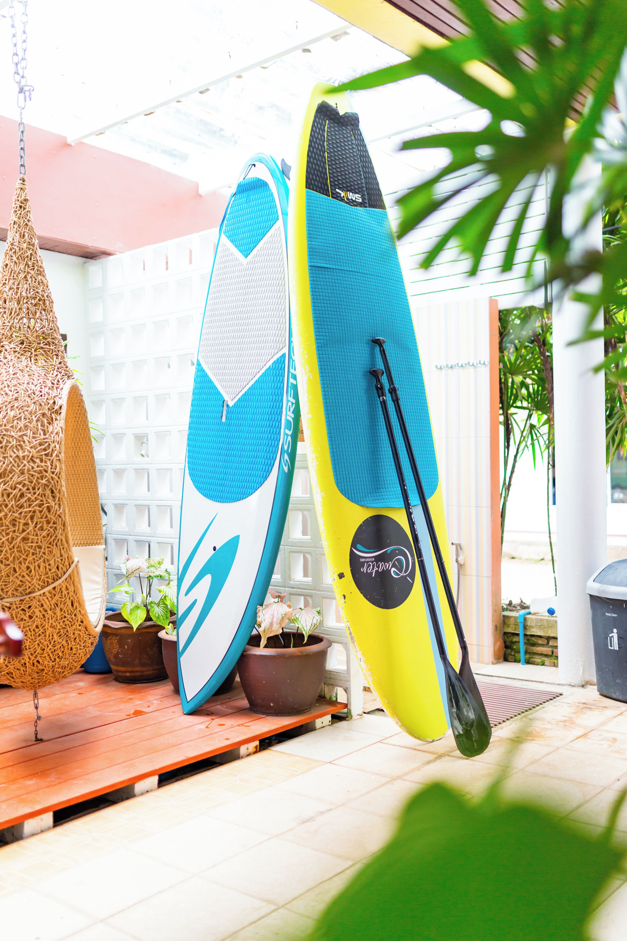 Two paddleboards leaning against a wall, surrounded by potted plants and a wooden deck.