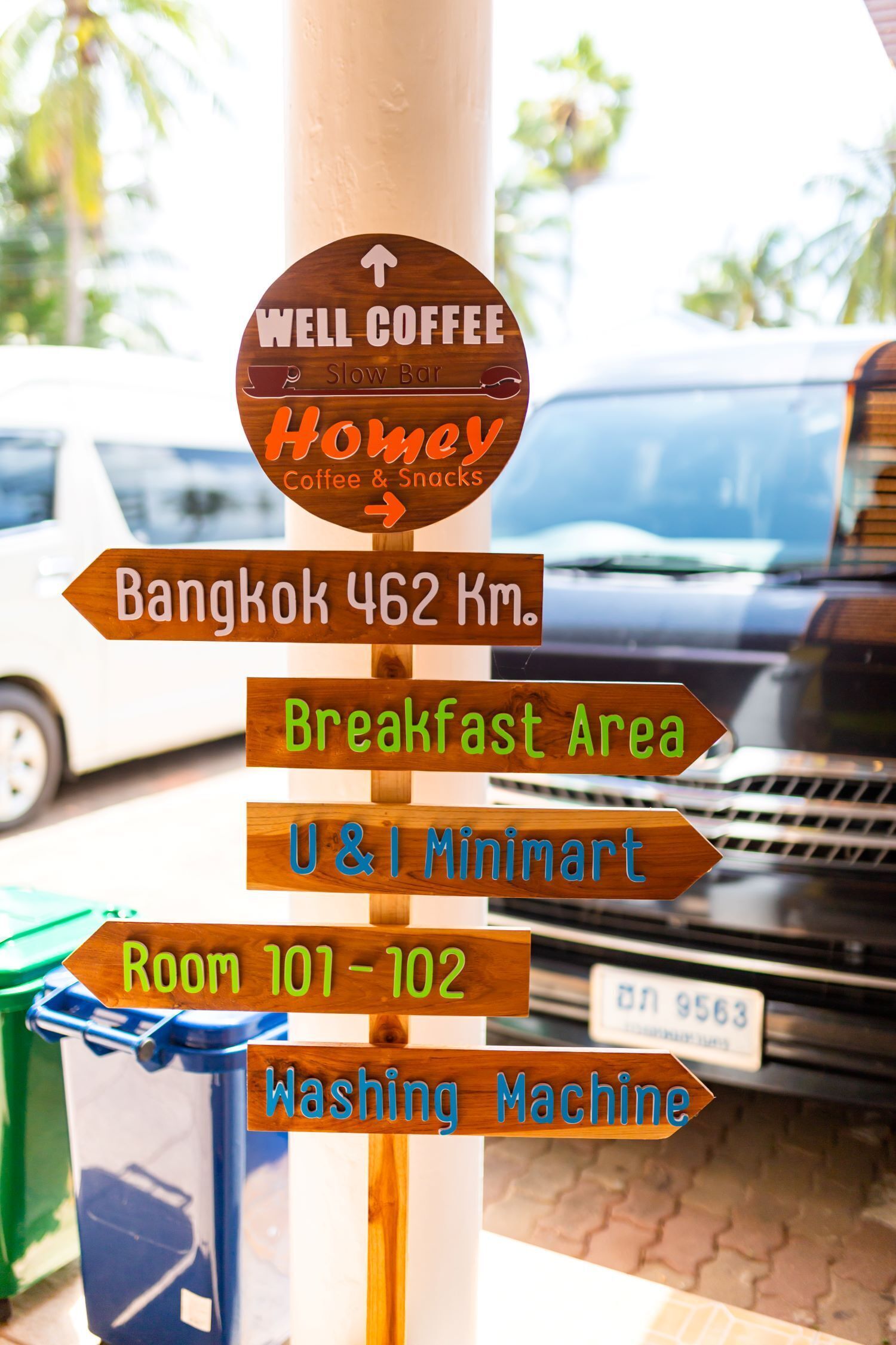 Un cartel direccional con múltiples flechas indicando varios lugares como Well Coffee, Homav Coffee & Snacks, Bangkok a 462 Km, Área de Desayuno, U&I MiniMart, Habitaciones 101-102 y Lavandería.