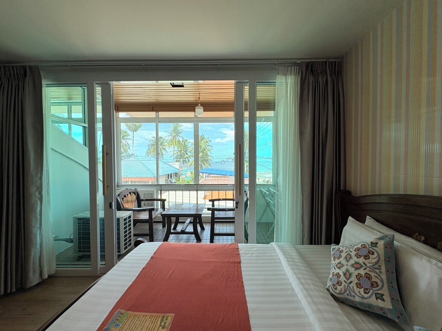 A hotel room with a bed, a table, and chairs outside the balcony, with a view of palm trees and buildings.