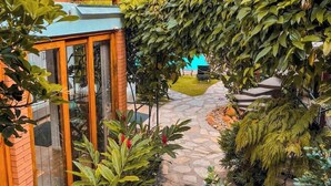 Um caminho de jardim leva a um deck de madeira com estrutura de vidro, cercado por vegetação exuberante e um caminho de pedra.