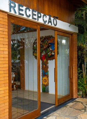 A reception area with a wooden facade, a glass door, and a colorful mural of birds and a sunflower. - Pousada Casa de Maria (Prado)