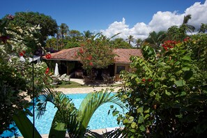 A pool with a tiled deck, surrounded by lush greenery and a building with a tiled roof. - Pousada Casa de Maria (Prado)