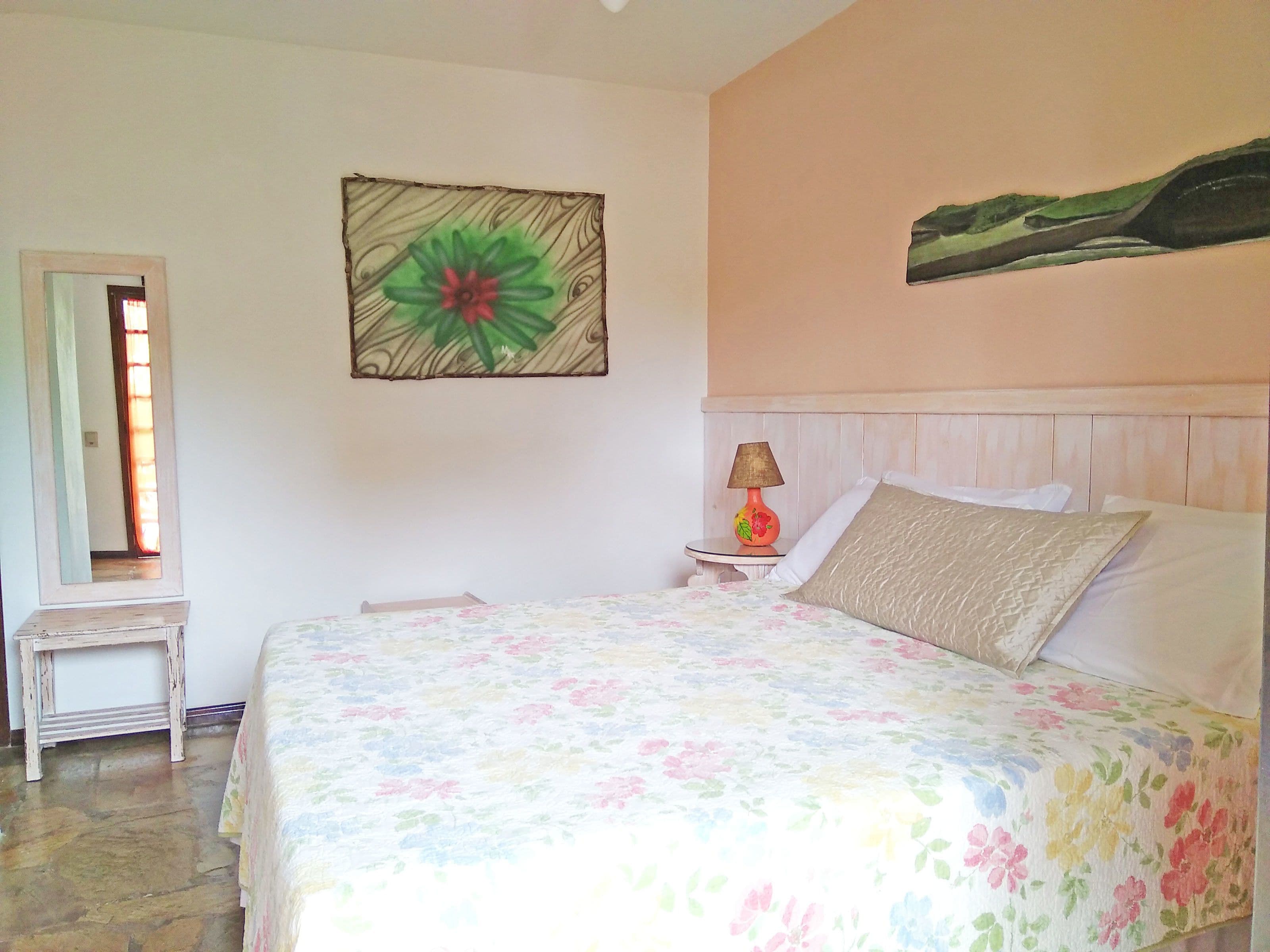 A bedroom with a bed, a nightstand, a lamp, a mirror, and a painting on the wall.