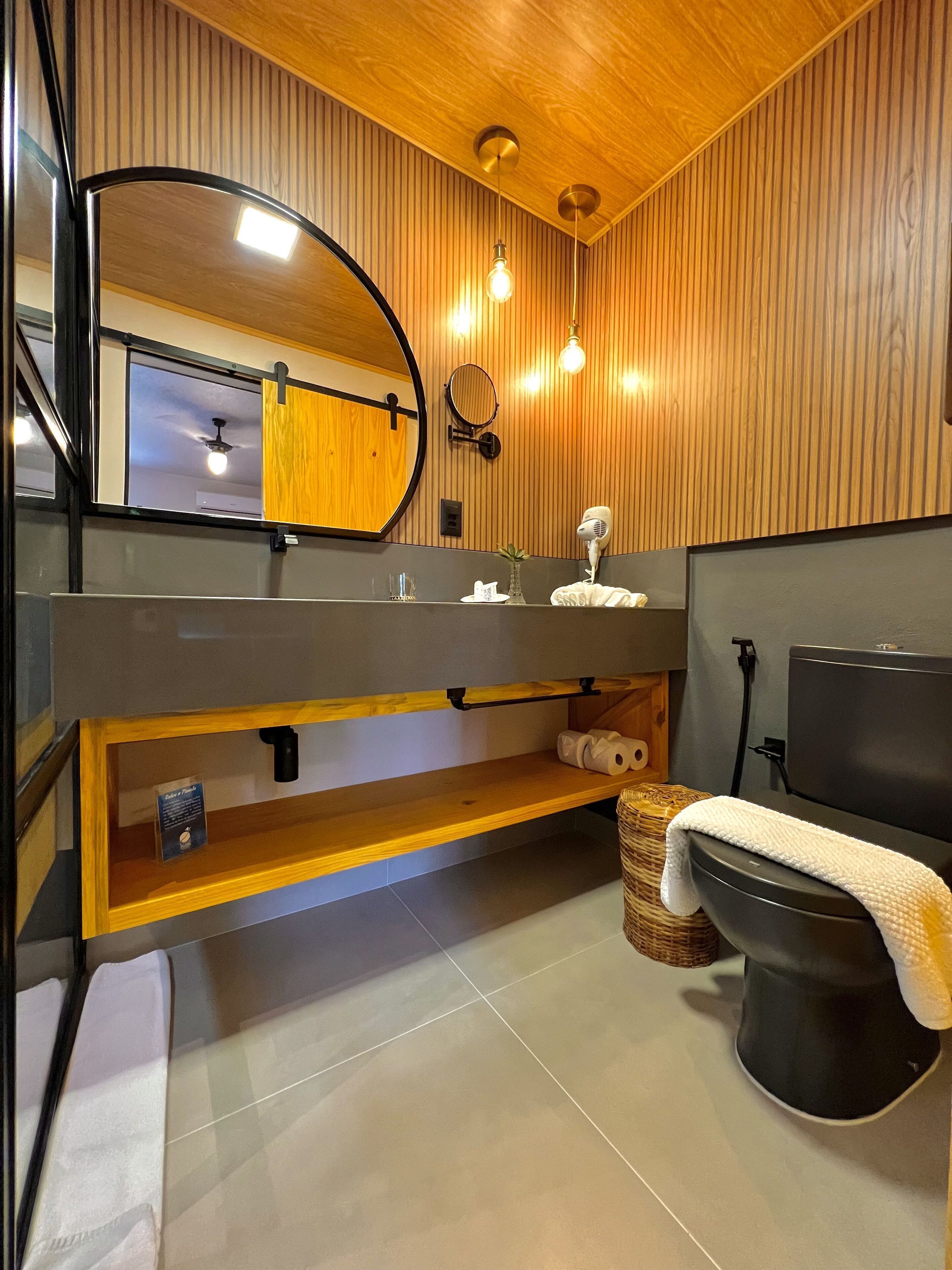 Un baño moderno con un tocador de madera, un espejo grande, un espejo redondo, una toalla, un inodoro y un pequeño estante con un cartel.