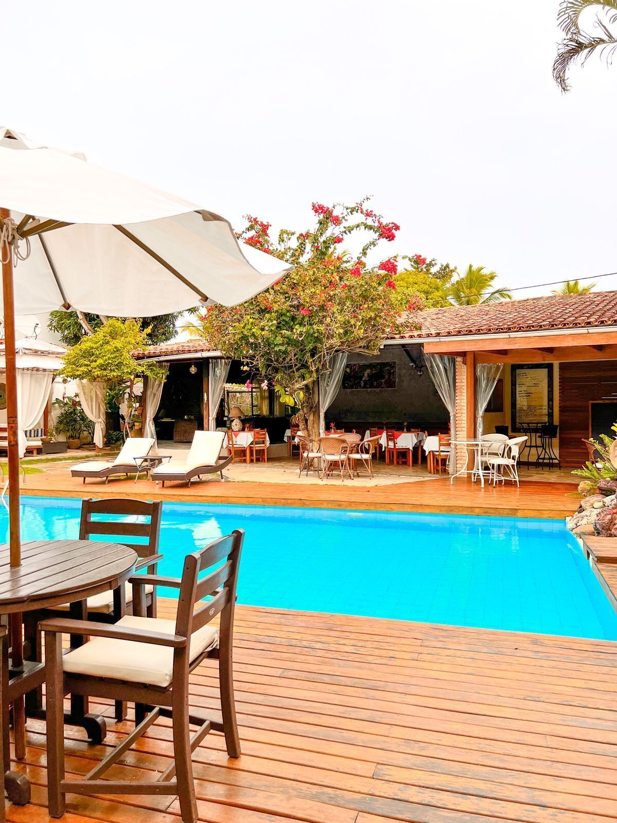 Un área junto a la piscina con sillones reclinables, una terraza de madera y una zona cubierta con mesas y sillas.