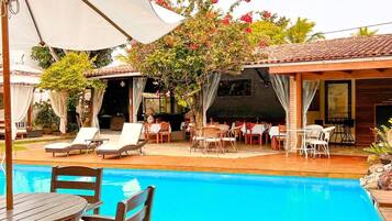 Un área junto a la piscina con sillones reclinables, una terraza de madera y una zona cubierta con mesas y sillas.
