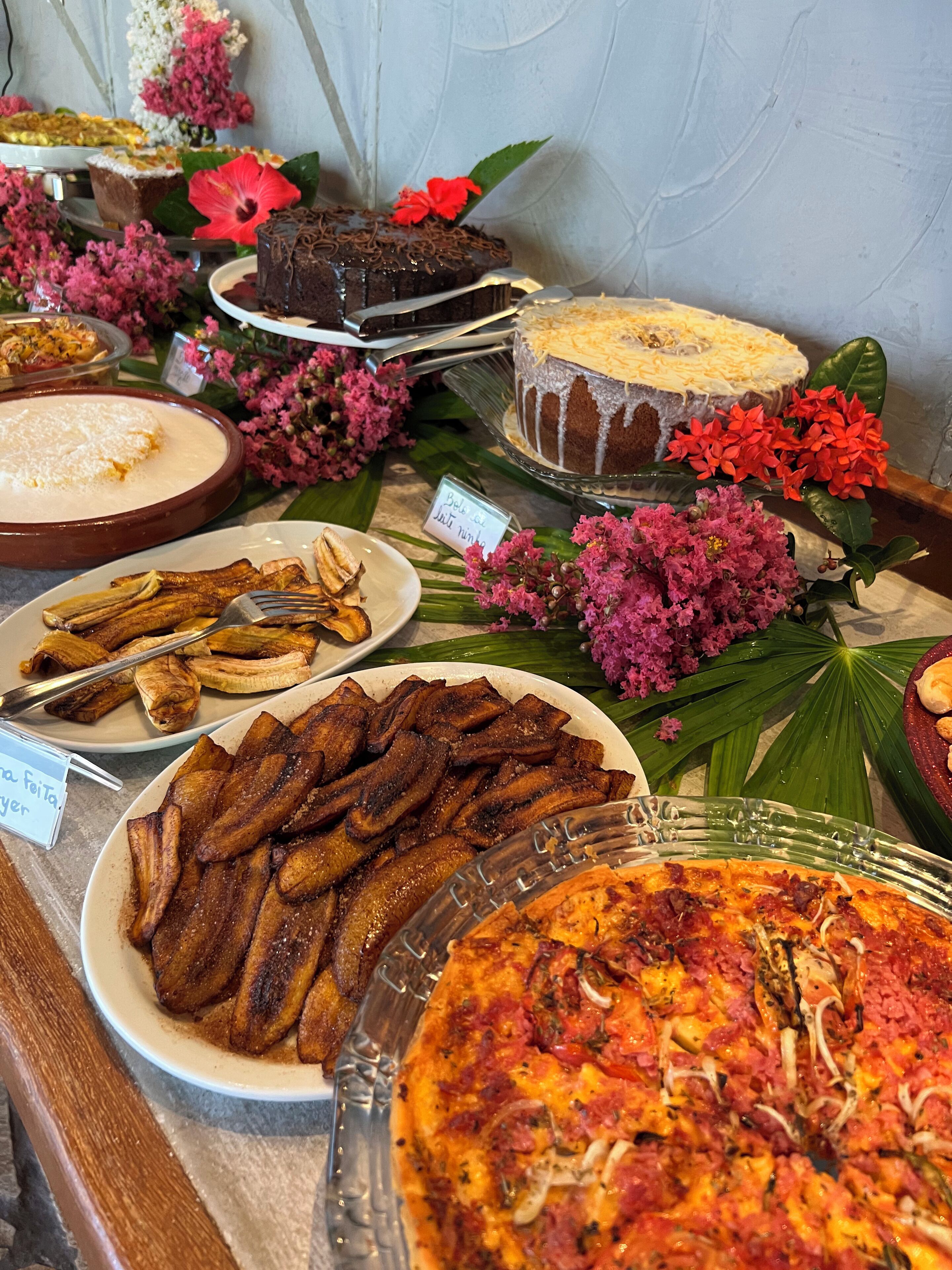 A buffet table with various dishes including a large pizza, fried plantains, and a chocolate cake.