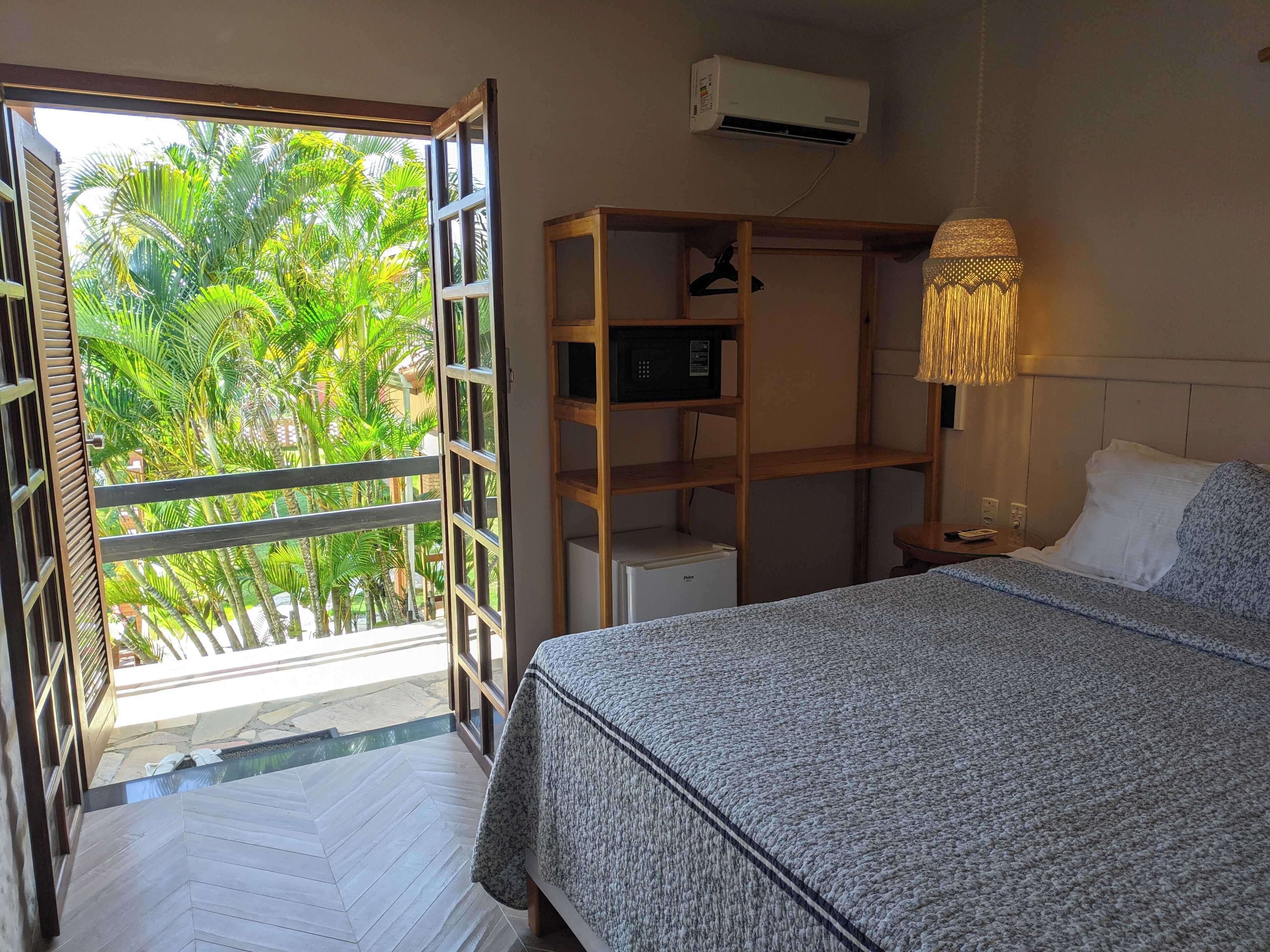 A hotel room with a bed, a wardrobe, a bedside table, an air conditioning unit, and a view of greenery through an open door.