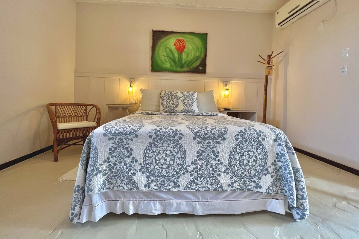 A bedroom with a large bed, two bedside tables, a chair, an air conditioner, and a painting on the wall.