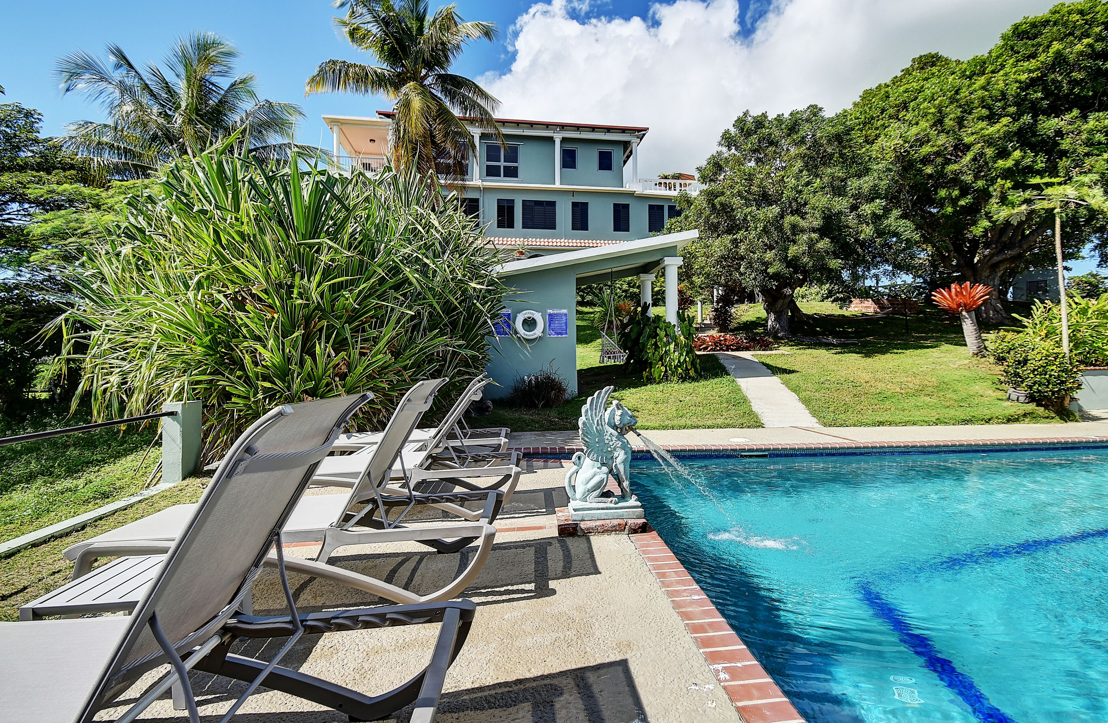 A pool area with lounge chairs and a statue, surrounded by greenery and a building in the background.