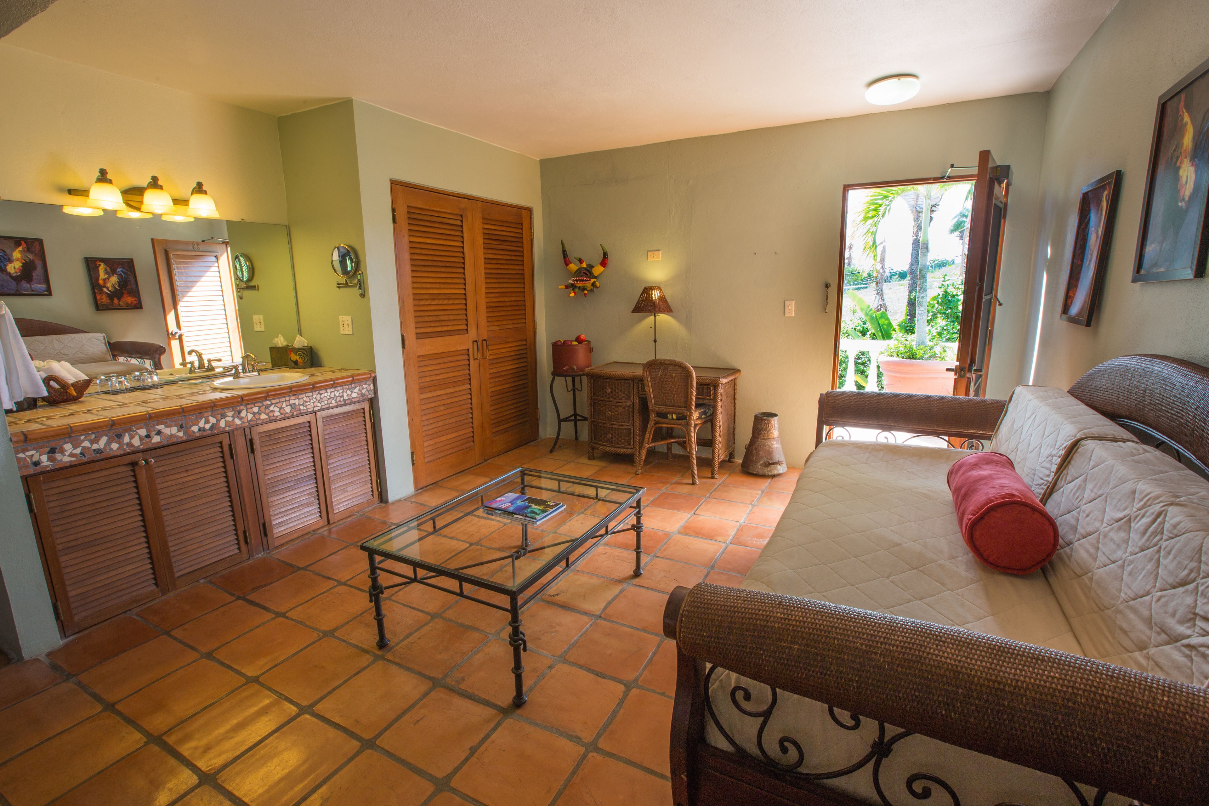 A room with a bed, a dining table, a chair, a small table with a book, a mirror, a lamp, and a window with a view of palm trees.