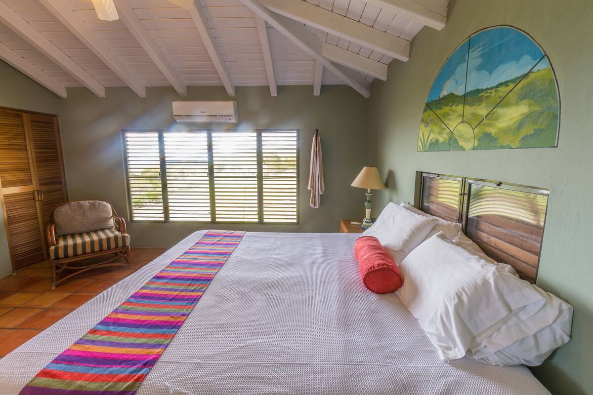 Une chambre spacieuse avec un grand lit, un tapis à rayures colorées, un fauteuil à bascule et une vue sur l’extérieur par la fenêtre.