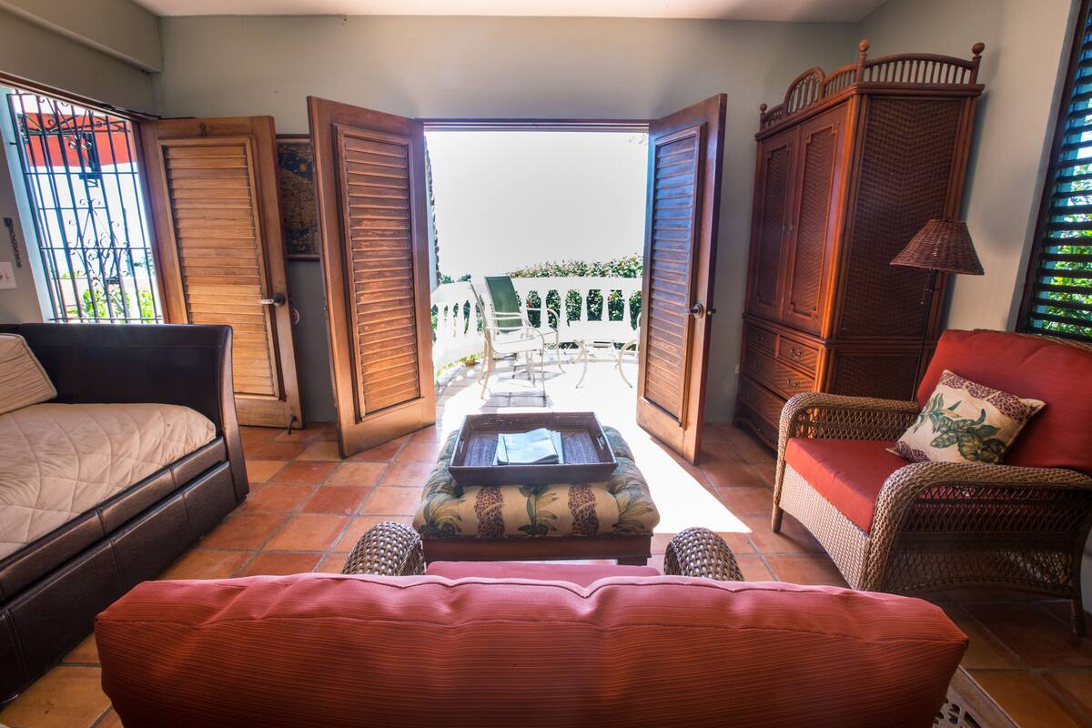 Une pièce comprenant un lit, un canapé rouge, des meubles en osier et donnant sur un balcon avec un espace salon extérieur.