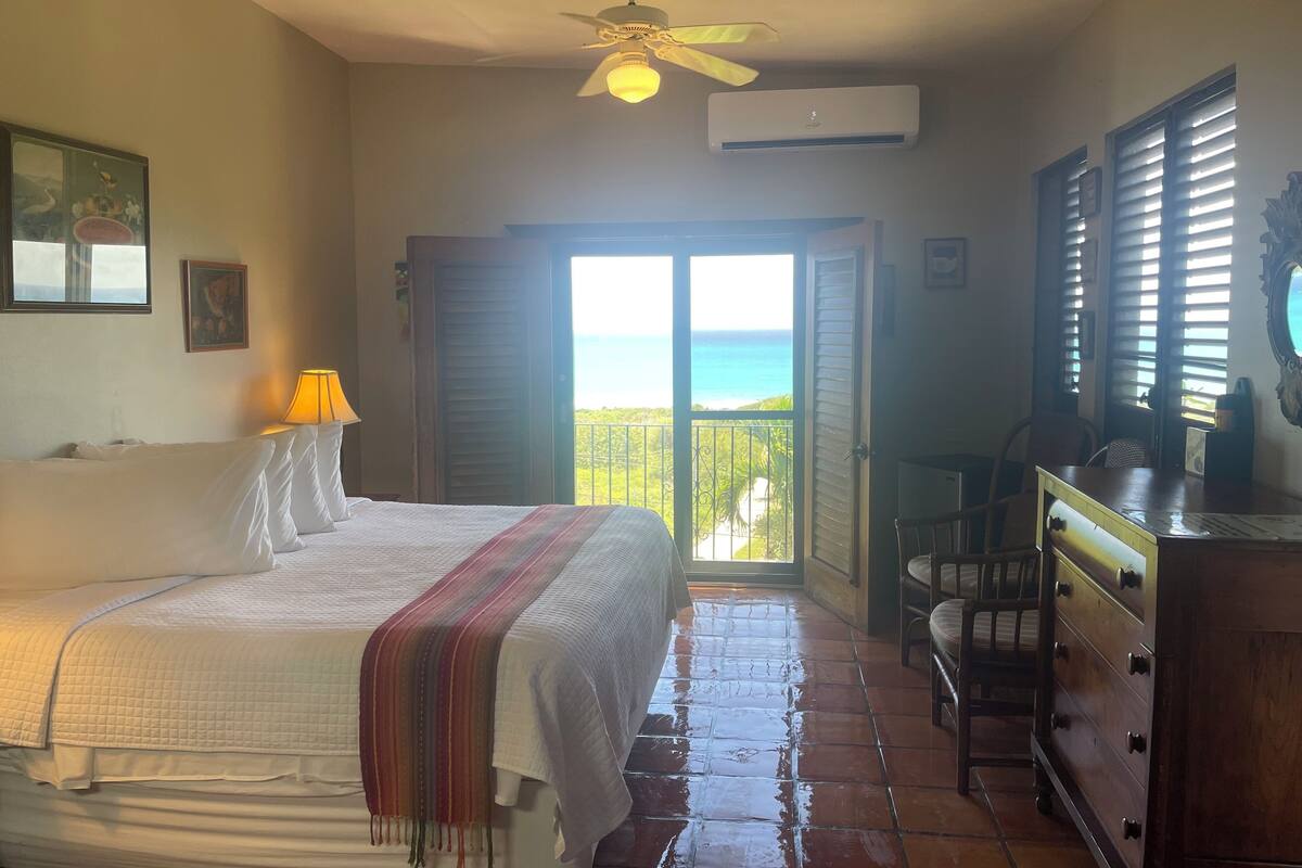 Une chambre à coucher comprenant un lit, une table de chevet, une lampe, une commode et une vue sur l’océan, visible à travers une porte coulissante.