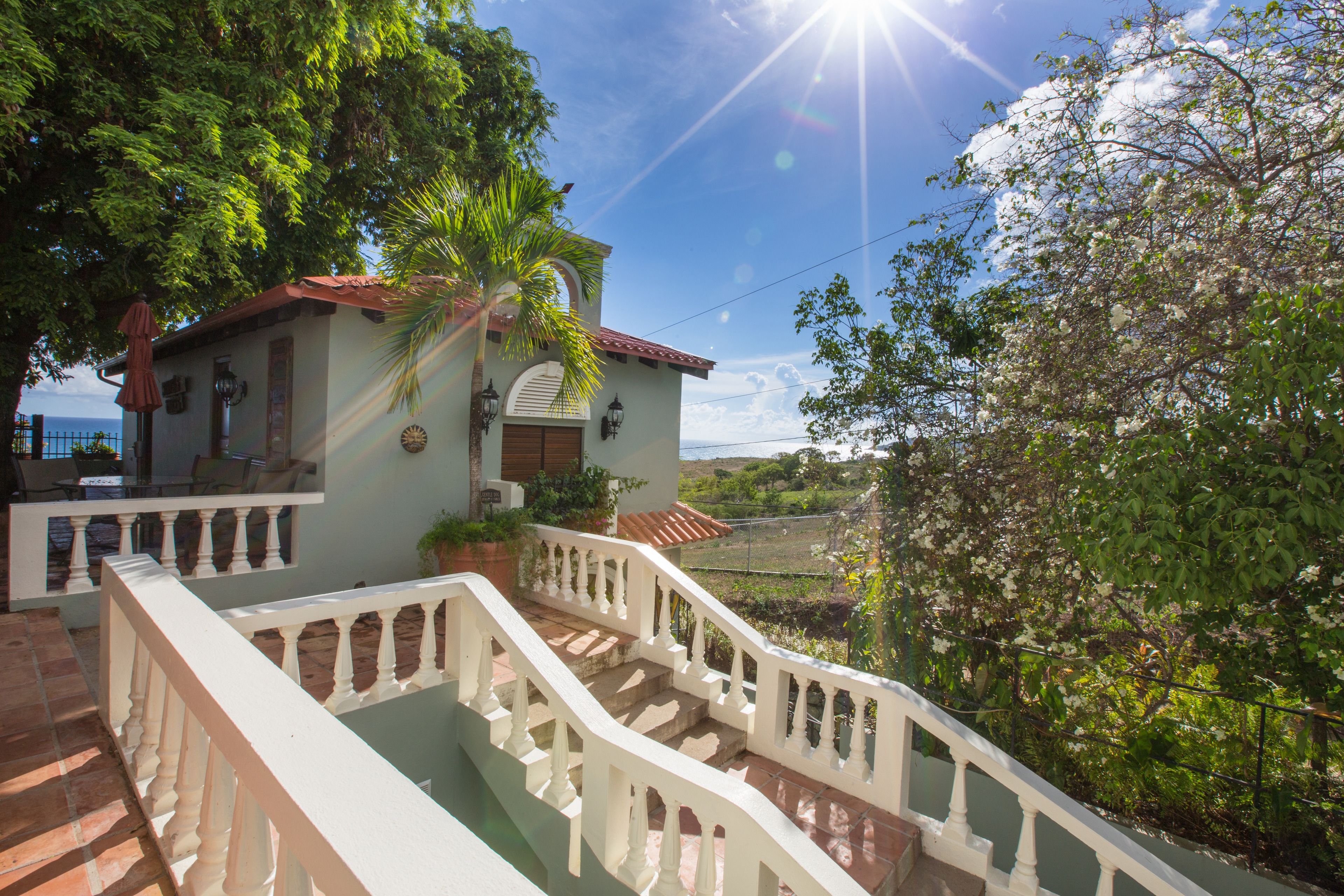 A house with a staircase leading to a balcony, surrounded by lush greenery and a clear sky.