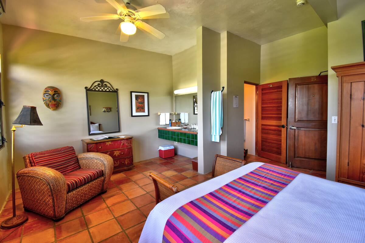 Une chambre d’hôtel comprenant un lit, un canapé, une chaise, une commode, un miroir, un ventilateur de plafond et une salle de bain visible à travers une porte ouverte.