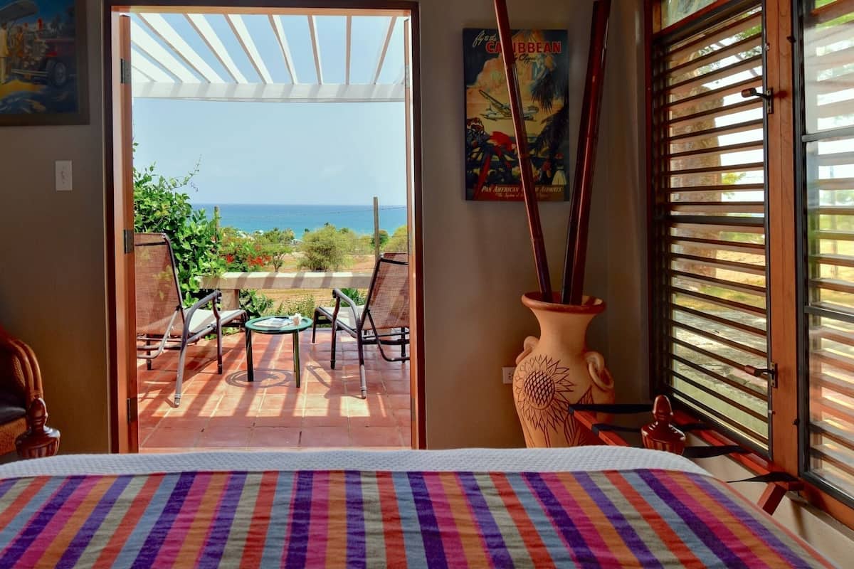 Une chambre à coucher avec un lit, une vue sur l’océan, une terrasse avec une table et des chaises, et un vase contenant des objets décoratifs.