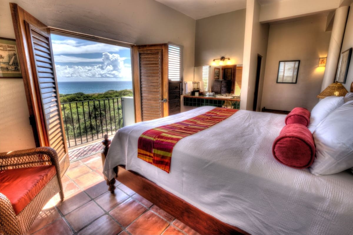 Une chambre avec un grand lit, un balcon avec vue sur l’océan, un bureau avec une chaise et une salle de bain avec un miroir et un lavabo.