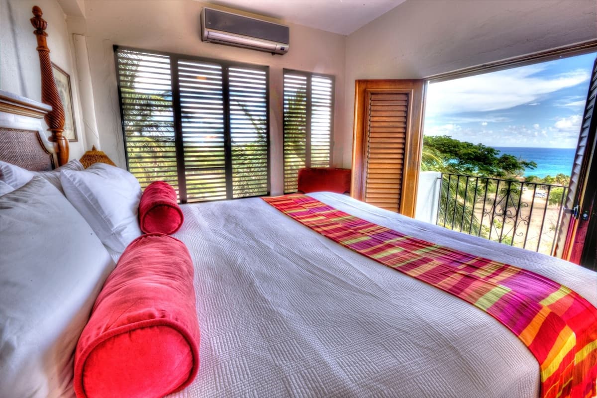 Une chambre d’hôtel avec un grand lit, un balcon donnant sur l’océan et une fenêtre équipée de stores.