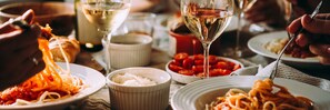Une table à manger avec différents plats, notamment des pâtes et un bol de riz, ainsi que plusieurs verres de vin blanc.