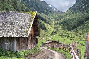 A rustic wooden building with a sloped roof, a dirt path leading to a fenced area, and a valley with greenery and a winding road in the background. - Appartments Am Bach (Gerlos)