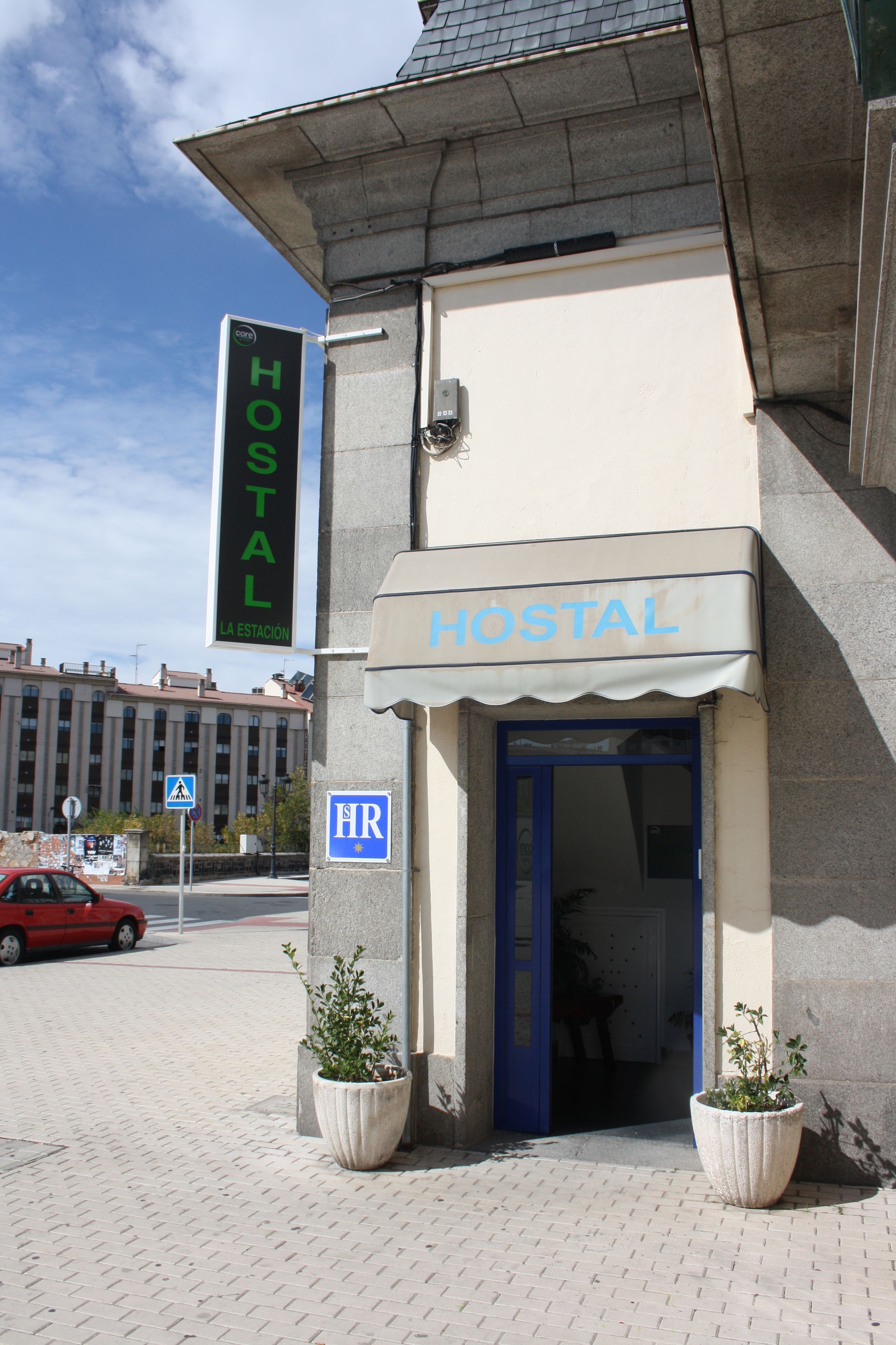 Un edificio con un letrero que dice "HOSTAL" y una puerta azul, que indica que es un albergue. Hay dos plantas en macetas al frente.
