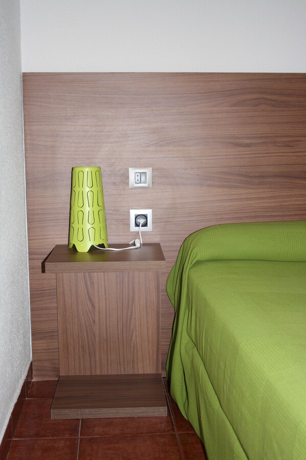 Habitación de hotel con cabecera de madera, edredón verde, lámpara verde sobre una mesita de noche de madera y dos enchufes.